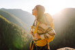 © maxbelchenko - A female hiker with a bright yellow backpack sits on a cliff and looks at the mountain range. Young woman feels freedom and enjoys the mountain landscape at sunset. Freedom and adventure concept.