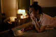 © m-art - A young woman with glasses is lying on her stomach in bed, focused on her laptop in a dimly lit room. Soft light from a nearby lamp creates a cozy atmosphere.