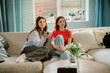 © m-art - Two young women sit comfortably on a sofa in a bright living room. One holds a remote, while the other looks thoughtfully at the screen. Afternoon light filters through the window.