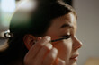 © m-art - A young woman carefully applies mascara to her eyelashes while sitting in a bright room. Soft sunlight filters through a window, enhancing her focused expression and room ambiance.