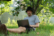 © Mihail - A man sits on the grass under a tree, focused on his laptop while contemplating. Surrounding greenery enhances the tranquil work environment in nature.