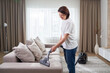 © Maria - young woman in white shirt and jeans cleaning carpet under sofa with vacuum cleaner in living room, copy space. Housework, cleanig and chores concept