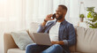 © Prostock-studio - Happy young african american bearded man typing on computer and call by phone on sofa in living room interior. Communication remotely, success business at home, work, new normal and social distance
