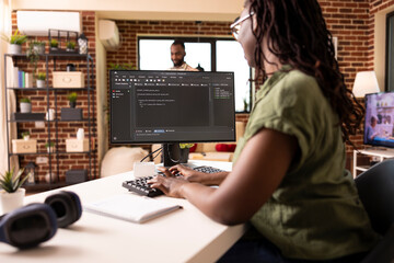 Remote female programmer works from cozy home desk, coding for startup project on desktop pc. Tech savvy black woman managing web development tasks on computer in brick wall apartment.