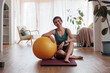© Jovo Jovanovic/Stocksy - Woman smiles with exercise ball on yoga mat at home.