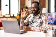 © DC Studio - African American male freelancer gestures hello on video call, attending remote skills workshop. Friendly man with headphones greets remote colleagues during virtual training session.