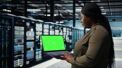 Programmer in data center using app on isolated screen laptop to ensure error free mainframes networks. Worker uses chroma key notebook to monitor server hub gear parts for failures, camera A