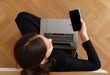 © muh - Overhead shot of a woman using a laptop and holding a phone. She's in black clothing, relaxing on a light-wood floor with neutral tones
