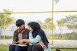 © Fajar - Two diverse university students collaborating on a project outdoors, with a Muslim woman in a hijab guiding her male classmate as they study together