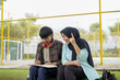 © Fajar - A moment of inspiration between two diverse Asian college students studying outdoors. A smiling young woman in a hijab explains her creative idea to a male classmate writing in a notebook