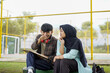 © Fajar - A happy young man and a Muslim woman in a hijab sitting together outdoors, collaborating on a school project and sharing ideas while looking at a digital tablet
