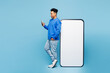 © ViDi Studio - Full body side view young man of African American ethnicity in sweatshirt casual clothes stand near big huge blank screen mobile cell phone with area use smartphone isolated on plain blue background