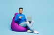 © ViDi Studio - Full body young smart IT man wear sweatshirt casual clothes sit in bag chair hold use work point on laptop pc computer isolated on plain pastel blue cyan background studio portrait. Lifestyle concept.