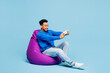 © ViDi Studio - Full body young man of African American ethnicity wears sweatshirt clothes sit in bag chair use play racing app on mobile cell phone hold gadget for pc video games isolated on plain blue background.