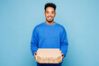 © ViDi Studio - Young man of African American ethnicity in sweatshirt casual clothes hold pizza in cardboard flatbox look camera isolated on plain pastel light blue cyan background studio portrait Lifestyle concept