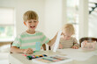 © m-art - Two children are engaged in creative art activities at a table. A boy smiles widely while holding a piece of paper, and a girl concentrates on her drawing. Sunlight fills the room.