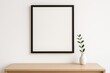 © CrazyJuke - Frame and a touch of Nature: Minimalist aesthetic composition featuring an empty black frame hung above a simple wooden surface adorned with an elegant vase holding a sprig of greenery.