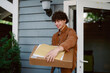 © m-art - A young man with curly hair is happily accepting a package at his front door. He is dressed casually and standing in a welcoming home environment during daylight.