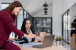 © MINAE - Two asian businesswomen are analyzing financial charts and data on a laptop and tablet, discussing and collaborating on a project in a modern office while a photographer takes pictures of them