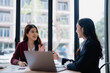 © MINAE - Two asian businesswomen are discussing strategy during meeting in modern office using laptop and financial documents, smiling and collaborating effectively