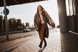 © deagreez - Young woman walking on an urban sidewalk in autumn, wearing a stylish coat, enjoying sunlight and expressing carefree elegance.
