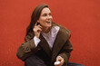 © zvkate - Happy laughing woman listen music, talk by phone using wireless headphones, she squatting. Woman wear raincoat short trench and long sleeves. Brunette long hair woman on orange background.