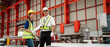 © NewSaetiew - Two industrial workers wearing safety helmets and reflective vests discussing checklist inspection in a modern metal sheet factory, focusing on teamwork, safety, and quality control in production.