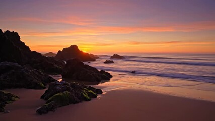 Wall Mural - Majestic Coastal Sunset with Rocks - A breathtaking sunset paints the sky with vibrant oranges and purples, casting a warm glow on a sandy beach dotted with dark rocks covered in green moss.
