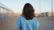 © zeenika - Female inmate standing alone in outdoor prison yard wearing blue uniform facing barbed wire fence under evening light.