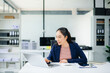 © laddawan - Confident Asian businesswoman using tablet in modern office with laptop and documents, showcasing finance, remote work