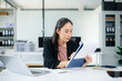 © laddawan - Smiling Asian businesswoman texting on phone at modern office desk with laptop, coffee, and documents