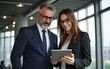 © fu - Team of mature senior leader man in suit and young employee woman discussing project on tablet standing in office. Two colleagues of professional business people looking at screen device. Vertical