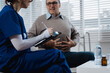 © NanSan - A young Caucasian nurse provides medical consultation to an elderly Caucasian man sitting on a sofa, addressing chronic conditions like obesity, hypertension, diabetes,heart disease, dementia, joint