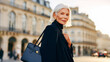 © dinatychynska - Stylish mature woman walks confidently through European city square. Elegant senior lady in black coat carries luxury handbag. Professional portrait showcases graceful aging and sophisticated style.