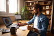 © Stockphotodirectors - A man in a casual outfit is engaged in work at a home office, analyzing data on multiple screens and taking notes, surrounded by books and plants, in a bright and inviting space.