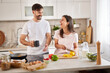 © Stockphotodirectors - A couple is happily interacting in a well-lit kitchen. One partner holds a mug, while the other chops vegetables on a cutting board, creating a joyful atmosphere.