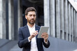 © Liubomir - Bearded businessman standing confidently in front of an office building, using a digital tablet to work remotely, showcasing modern technology and efficient communication in an urban setting