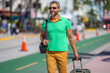 A tourist walks through the summer city streets. A man enjoys his summer vacation in a tropical summer city. Traveling with a suitcase. A full-length portrait of a traveler on an urban summer
