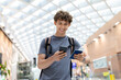 © Anastasiya - Young Caucasian man with backpack holding passport and smartphone, smiling inside airport terminal. Travel concept of departure, boarding, youth lifestyle and digital routine