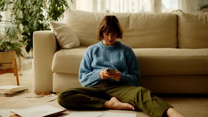 Serious woman typing text message on her smartphone while sitting on the floor in the living room