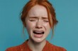 © Cheewynn - A young woman with red hair is crying with her eyes closed against a blue background, showing a strong emotional reaction