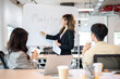 © bongkarn - Asian businesswoman standing writing on whiteboard explaining marketing plan to coworkers in office.
