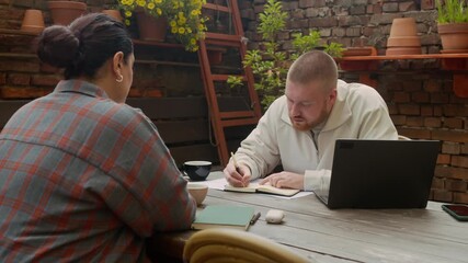 Wall Mural - Medium long shot of two intercultural colleagues sitting at table with wireless laptop and documents, taking notes and drinking coffee while discussing business plan in outdoor cafe