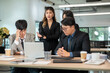 © bongkarn - Serious businesswoman frown talking while man coworkers looking at laptop on wooden table in office.