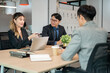 © bongkarn - Businesswoman holding pen talking over laptop sitting with coworkers at wooden table in meeting room