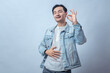 © Daniel - Asian man in denim jacket smiling with eyes closed, touching stomach with one hand and showing thumbs up, standing in studio against plain light background, expressing relief and satisfaction