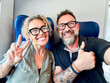 © simona - Middle-aged couple taking selfie together while sitting on train, countryside landscape visible through window, smiling and happy, enjoying travel, love, leisure, adventure and modern lifestyle