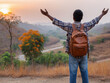 © Gesfera Images - Energetic backpacker raising arms, celebrating mountain hike achievement with vibrant sunset backdrop
