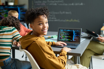 Portrait of Black teenager boy smiling and turning toward camera while coding on laptop in classroom, classmates working at desks in background, chalkboard with programming notes visible