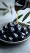 © Yuliia - Fresh black olives on ceramic plate with golden olive oil pouring from bottle, green leaves in background. Close up food photography on gray surface.